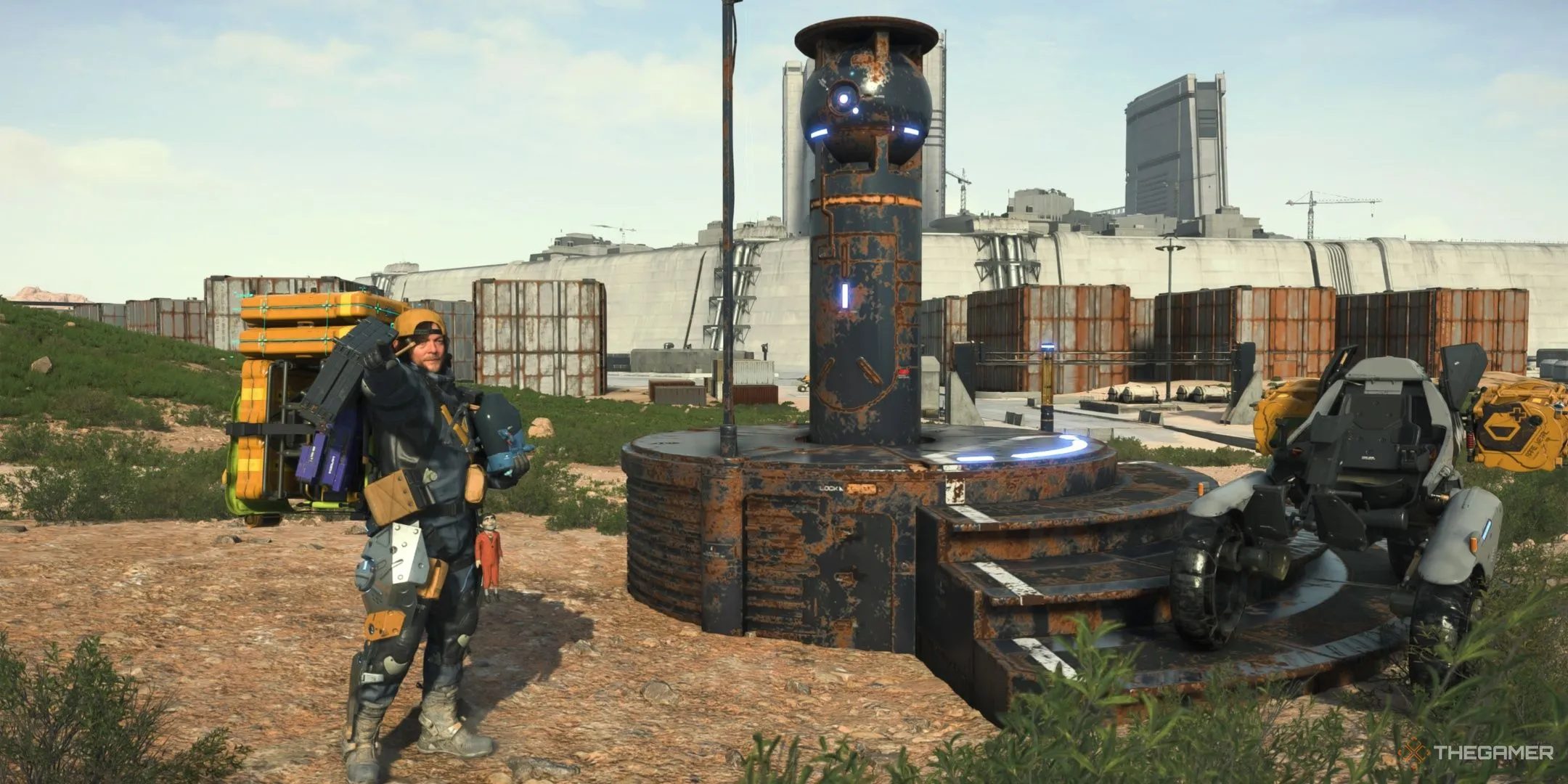 Sam Porter Bridges tạo dáng cùng BB pod trước một máy lát đường tự động bị hỏng trong Death Stranding 2: On the Beach, nhấn mạnh yếu tố phiêu lưu và giao hàng.